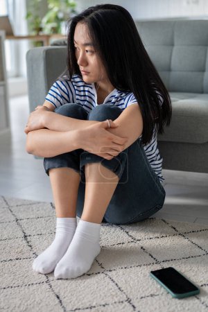 Photo for Worried anxious Asian Chinese woman feeling upset abandoned after quarrel waiting call or message sits on floor hold knees at home. Distressed Korean girl lost in thoughts about relationship troubles. - Royalty Free Image