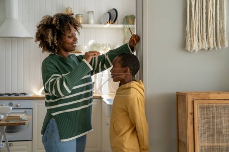 Photo for African American stay-at-home mom measuring height of kid son at home, school-aged boy standing against wall while mother taking measurement with ruler and pencil. Child growth and development concept - Royalty Free Image
