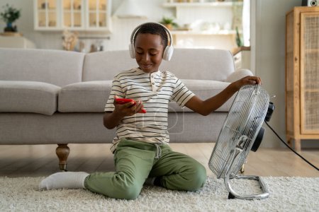 Foto de Feliz niño afroamericano escolar disfrutando de tiempo libre después de la escuela en casa, sentado en el suelo frente a un ventilador eléctrico, sosteniendo el teléfono inteligente jugando juego en línea. Calor de verano y niños - Imagen libre de derechos