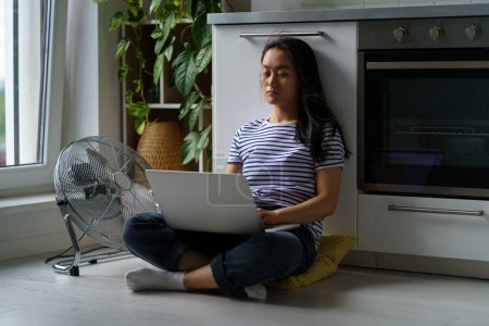 Photo for Tired Asian woman sitting on kitchen floor in front of ventilator. Exhausted Chinese girl suffering from high temperature at home by hot weather, cooling with air conditioner while work with laptop. - Royalty Free Image