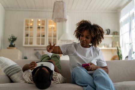 Téléchargez les photos : Enfant afro-américain jouant à des jeux VR tout en passant du temps avec maman à la maison. Famille moderne, maman et enfant se détendre sur le canapé avec des appareils. Dangers de la réalité virtuelle pour les jeunes enfants - en image libre de droit