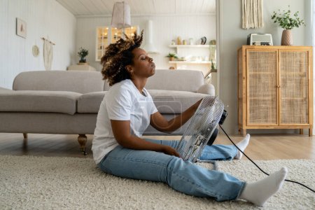 Téléchargez les photos : Fatiguée femme noire surchauffée se refroidissant avec un ventilateur électrique à la maison, souffrant de chaleur tout en étant assise sur le sol dans le salon sans climatiseur, gardant au frais à l'intérieur pendant la canicule d'été - en image libre de droit