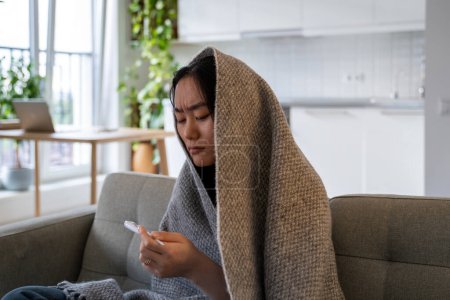 Téléchargez les photos : Mauvaise santé asiatique chinoise jeune femme assise sous une couverture de laine sur un canapé souffrant de froid, regardant thermomètre mal à l'aise avec la fièvre élevée maladie du virus de la grippe migraine de grippe froide. - en image libre de droit