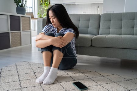 Worried anxious Asian Chinese woman feeling upset abandoned after quarrel waiting call or message sits on floor hold knees at home. Distressed Korean girl lost in thoughts about relationship troubles.