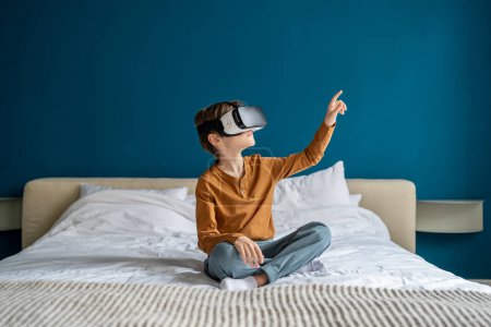 Téléchargez les photos : Enfant garçon portant casque de réalité virtuelle interagissant avec le monde numérique, enfant curieux explorer l'univers avec vr, assis sur le lit à la maison. Enfants et expérience d'apprentissage immersive, métaverse pour les enfants - en image libre de droit