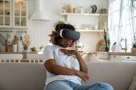 Téléchargez les photos : Effrayée surprise femme afro-américaine porter casque de réalité virtuelle immergé dans métaverse jeu vidéo regarder film d'horreur se trouve dans la pose ferme de la peur. Femme noire choquée dans des lunettes 3D à la maison - en image libre de droit