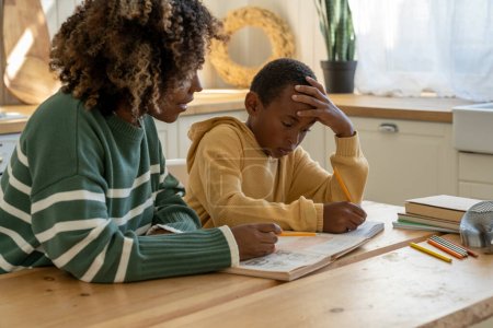 Homeschooling together. Caring African American mother helping thoughtful adopted son with tasks of school homework for effective education. Attentive private black tutor woman give lesson pensive boy