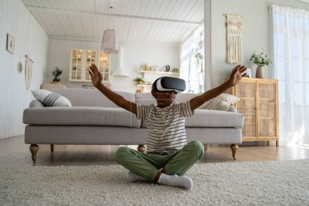 Téléchargez les photos : Enfant numérique. Petit garçon afro-américain jouant au jeu de simulation VR, écolier curieux explorant les planètes et les étoiles en réalité virtuelle et augmentée, assis sur le sol et touchant l'air avec les paumes - en image libre de droit