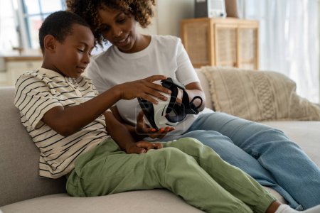 Téléchargez les photos : Femme afro-américaine satisfaite tient des lunettes de réalité virtuelle pour smartphone avec un petit fils heureux. Joyeuse maman noire show écolier VR casque pour 360 métaverse de jeu d'immersion vidéo à la maison - en image libre de droit