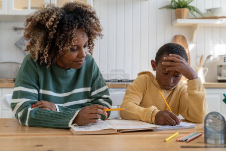 Photo for Focused biracial child boy looks at study book while listening to African American tutor female at private home lesson. Educational work to focused kid son with mom helping with homework assignment - Royalty Free Image