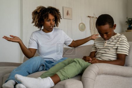 Photo for Dissatisfied African American woman mother scolding upset son for bad behavior while sitting together on sofa in living room. Disappointed parent mom talking with kid, disciplining child at home - Royalty Free Image