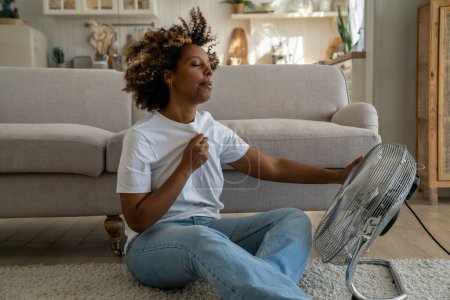 Photo for Summer heat. Young African American woman cooling down by ventilator at home, feeling unwell with high temperature during hot weather, sitting on floor in front of electric fan during extreme heatwave - Royalty Free Image