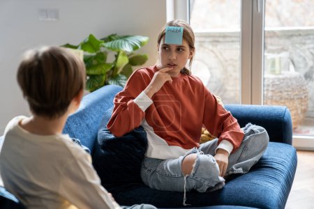 Photo for Thoughtful teenage girl guessing word on sticker on forehead while playing in who am i with younger brother sitting on couch. Interested playful older sister think about game note with friend at home - Royalty Free Image