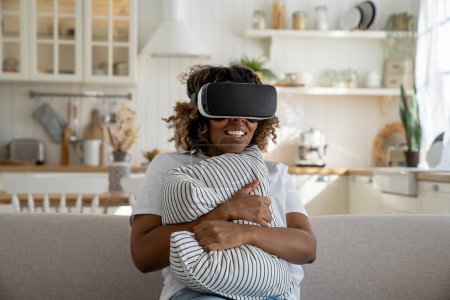 Téléchargez les photos : Femme noire excitée portant le casque VR assis sur le canapé tenant l'oreiller regardant passionnant film 3D fascinant, passer du temps libre dans la réalité augmentée. Réalité augmentée et divertissement à domicile - en image libre de droit