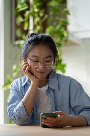 Foto de Buena mujer coreana asiática satisfecha con gafas elegantes mira en el teléfono móvil, lee buenas noticias chats con amigos disfruta de ir de compras en línea, mientras que el descanso se sienta en casa con las plantas después del trabajo o el estudio. - Imagen libre de derechos