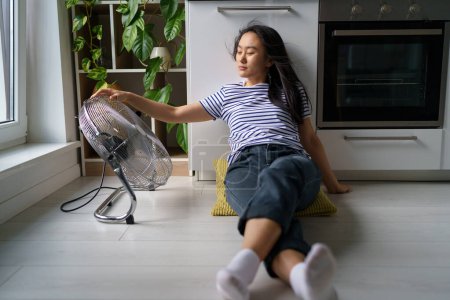 Photo for Overheating in homes. Tired overheated young Asian woman sitting on floor in kitchen near electric fan, cooling down at home during extreme summer heat, staying cool without air conditioning - Royalty Free Image