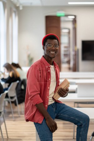 Photo for Education and happiness. Portrait of young cheerful happy African guy university student wearing glasses with pile of books in hands sitting on desk in classroom and smiling at camera, selective focus - Royalty Free Image