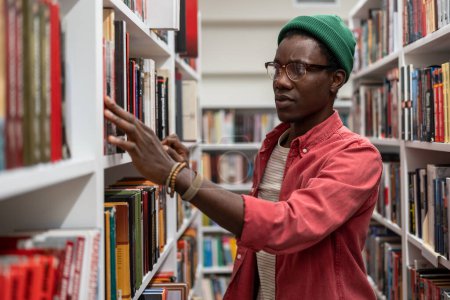 Téléchargez les photos : Étudiant africain de l'Université portant des lunettes debout entre les étagères de la bibliothèque, ramasser un livre sur une étagère, à la recherche de littérature tout en préparant un projet de recherche. Éducation et lecture - en image libre de droit