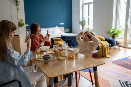 Photo for Playful boy covering eyes with bread toasts posing for photo, having fun while eating breakfast together with family members. People using smartphones during mealtime. Mobile devices and family time - Royalty Free Image