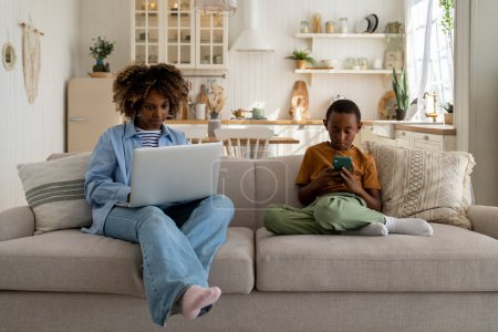 Photo for Focused working from home African mother freelancer using laptop while sitting on couch with child playing game on smartphone, woman mom freelancing with small kid. Motherhood and career concept - Royalty Free Image