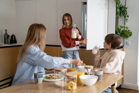 Photo for Family meal time. Single mother and two kids eating in kitchen at home, spending quality time together in morning. Smiling teen girl taking milk out of fridge while having breakfast with relatives - Royalty Free Image