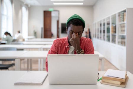 Foto de Estrés académico. Cansado hombre negro frustrado sentado en frente de la computadora portátil en la biblioteca de la universidad, que sufre de tensión ocular digital causada por la educación en línea. Síndrome de visión computarizada en estudiantes - Imagen libre de derechos