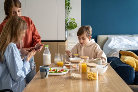 Foto de Madre e hija usando el teléfono móvil en la mesa de la cocina mientras desayunan junto con la familia. Chica adolescente mostrando a mamá algo en el teléfono inteligente durante la hora de la comida. Uso del teléfono celular y vínculo familiar - Imagen libre de derechos