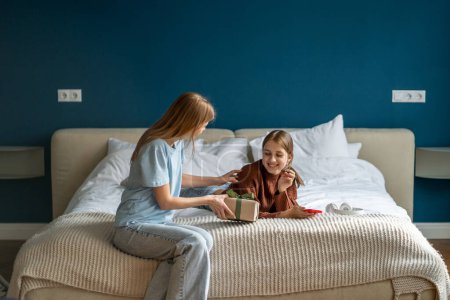 Photo for Happy surprised adolescent teen girl receiving birthday present from mother while resting on bed. Loving mom giving wrapped gift box to excited teenage daughter, congratulating child at home - Royalty Free Image