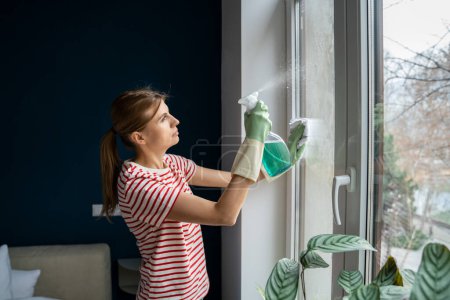 Photo for Attentive woman wash glass window wearing rubber protective gloves, with rag and eco spray bottle detergent. Focused professional maid cleaning hotel room. Housekeeping perfect tidiness domestic work - Royalty Free Image