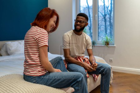 Photo for Communication in relationships. Young diverse interracial couple man and woman laughing while sitting on bed and talking, having good conversation during date night at home, bonding with partner - Royalty Free Image