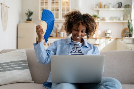Photo for Smiling satisfied African American woman waving blue fan in hand bring back good mood on hot day at home. Pleased black female escaping from heat stuffiness while online work with laptop sit on couch - Royalty Free Image