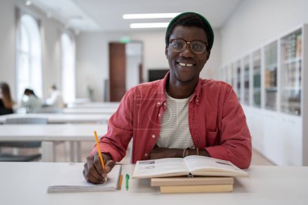 Téléchargez les photos : Étudiant afro-américain positif étudiant en bibliothèque prenant des notes dans un bloc-notes, souriant en regardant la caméra. Joyeux Noir se préparant pour les examens à l'université. Éducation, nouvelle profession. - en image libre de droit