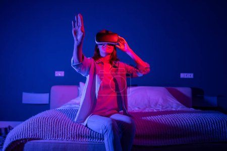 Photo for Joyful woman immersed in the metaverse in virtual reality glasses hands touches air interested while sit on bed in living room at night. Excited girl in 3D VR goggles at home with neon light. - Royalty Free Image