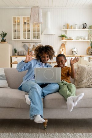 Téléchargez les photos : Heureuse mère afro-américaine joyeuse et son fils assis sur le canapé avec un ordinateur portable en agitant les mains et en regardant la caméra Web, avoir un appel vidéo avec la famille. Sourire maman et enfant garçon parler avec des parents en ligne - en image libre de droit