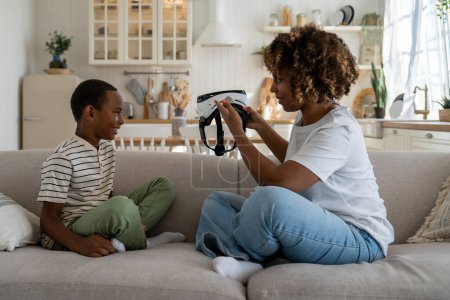 Téléchargez les photos : Jeune mère afro-américaine portant un casque VR introduisant la technologie de réalité virtuelle à l'enfant fils tout en passant du temps libre ensemble à la maison. Maman parent explorer le monde virtuel avec enfant - en image libre de droit