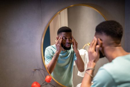 Photo for Worried african american man looks at mirror, touches facial skin anxious about forehead eyes wrinkles. Stressed serious guy has facial skin problems. Skincare, dermatology, face skin aging concept. - Royalty Free Image