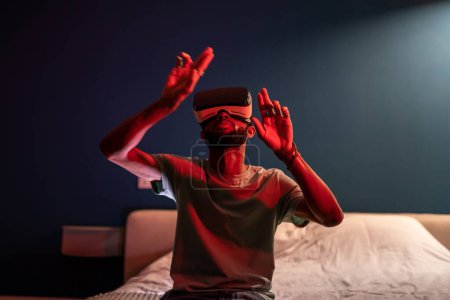 Foto de El hombre negro usa auriculares de realidad virtual interactuando con el mundo digital, curioso chico afroamericano explorando el universo con vr, jugando juegos 3D, sentado en la cama en casa con luz de neón. - Imagen libre de derechos