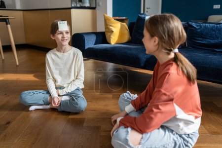 Photo for Smiling teen sister and child brother play together who am i guess sticker game sit on floor at home in living room. Girl has sticker with frog word on forehead. Family funny time, siblings friendship - Royalty Free Image