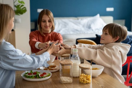 Photo for Loving mother with teen daughter and kid son sit at table hold warm cup together in breakfast. Happy fun family vow at kitchen table. Trusting relationship between parent and children be always team - Royalty Free Image
