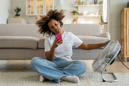 Photo for African American girl sitting on floor in living room near electric fan and using smartphone, unhappy black woman cooling down, trying to stay cool without air conditioning, suffering from heat - Royalty Free Image