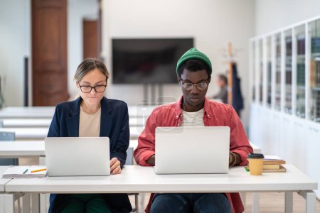Seriöse multiethnische Studenten an internationalen Universitäten arbeiten an Laptops in der Campus-Bibliothek sitzt am Schreibtisch. Afrikanischer Mann und kaukasische Frau studieren gemeinsam an Forschungsprojekt.