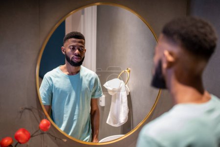 Téléchargez les photos : Malheureux bouleversé homme afro-américain de la trentaine regardant dans un miroir, debout dans la salle de bain à la maison, triste déprimé noir se sentant insatisfait de son apparence. Crise de la quarantaine chez les hommes - en image libre de droit