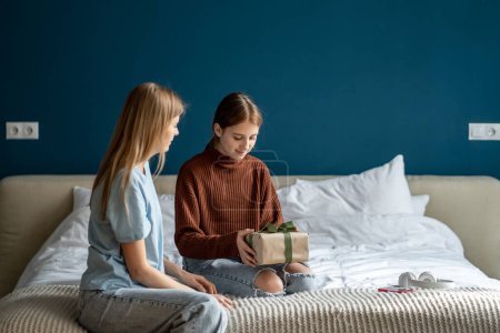 Photo for Mother congratulating teen girl daughter, giving wrapped gift box with ribbon. Caucasian family child and mom celebrate birthday at home together, adolescent kid sitting on bed opening present - Royalty Free Image