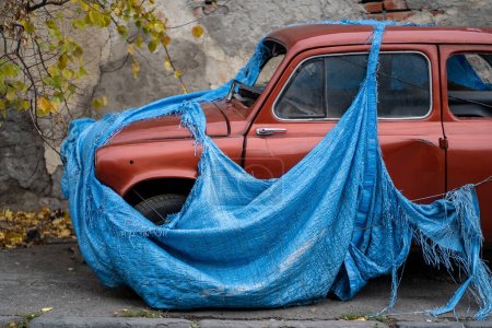 Photo for Retro old abandoned red car with broken glass covered with torn blue awning to prevent metal corrosion. Vintage small vehicle forgotten by owner stands on autumn street. unwanted discarded items - Royalty Free Image