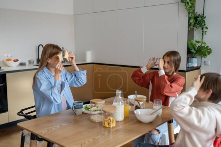 Téléchargez les photos : Joyeuse famille caucasienne jeune mère et deux enfants s'amusent pendant le petit déjeuner à la maison, se lient et apprécient le temps du matin ensemble à la maison. Maman profiter des premières heures avec les enfants avant l'école - en image libre de droit