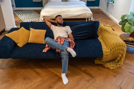 Photo for Tired pensive black man relaxing on comfortable couch enjoying lazy weekend at modern interior home. . Thoughtful bearded dreaming african american guy sits on sofa rests in studio apartment. - Royalty Free Image