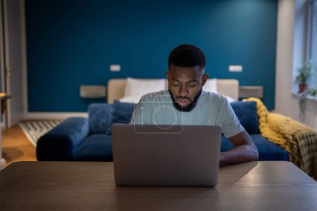 Photo for Young focused African American man freelancer sitting at table doing remote job on laptop until late hours, tired black guy working on project all night, using computer. Freelance and deadline - Royalty Free Image