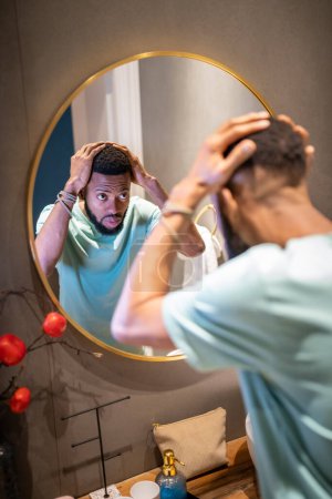 Téléchargez les photos : Hommes perte de cheveux. Jeune homme afro-américain bouleversé regardant dans la salle de bain miroir toucher les cheveux, mec noir déprimé s'inquiétant de la calvitie. Calvitie masculine et concept de santé mentale - en image libre de droit