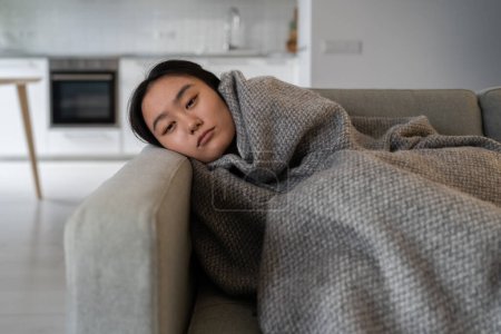 Photo for Sad tired Asian girl lying alone feels depression apathy indifference to everything melancholy attack has mental problems. Unhappy woman warming wrapped with plaid from central heating problem at home - Royalty Free Image