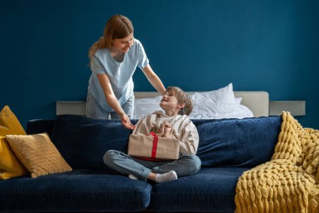 Photo for Happy little boy sitting on sofa with wrapped gift box, small child son feeling excited while getting birthday present from loving mother at home. Mom making holiday surprise to kid - Royalty Free Image